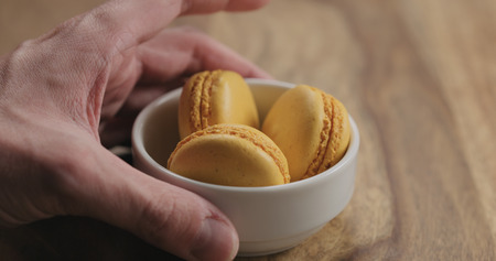 man hand put orange macarons in white bowl on wood table, wide photoの写真素材