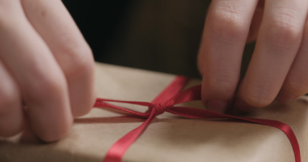 Macro shot of female hands tying red ribbon bow on craft paper gift box, wide photoの写真素材