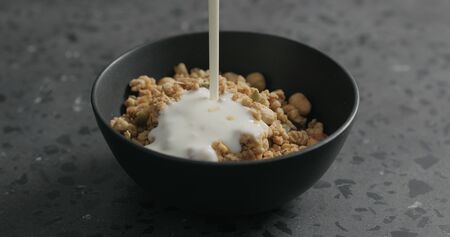 pour yogurt over whole grain granola with hazelnuts and pumpkin seeds in black bowl on terrazzo countertop, wide photoの写真素材