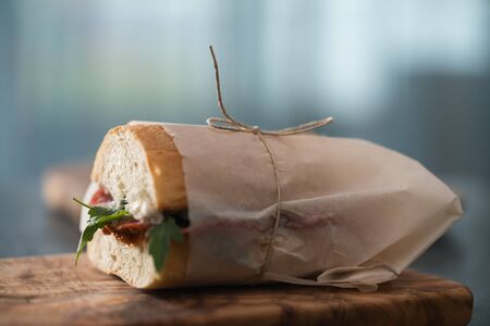 Ciabatta sandwich with salame, arugula, red pesto and cream cheese wrapped in paper on olive boardの写真素材