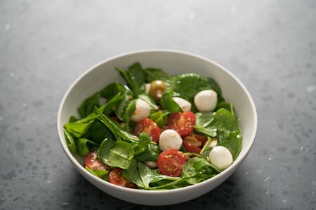 Salad with cherry tomatoes, spinach leaves and mozzarella balls in white bowl on terrazzo surface, shallow focusの写真素材