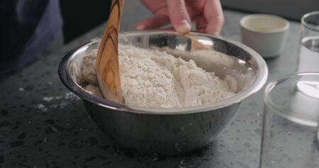 man mixing dry ingredients with flour in steel bowl on concrete countertop, wide photo