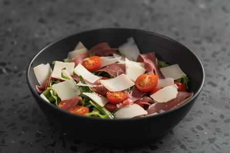 Salad with prosciutto, tomato, arugula leaves and parmesan cheese in black bowl on concrete surface, shallow focusの写真素材