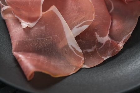 Thin italian prosciutto slices on black plate on conctere background closeup, shallow focus
