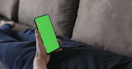 Closeup young man lying on a couch and holding smartphone with green screen, wide photoの写真素材