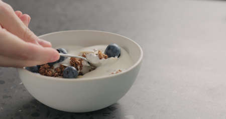 man hand tastes chocolate granola with almond flakes and blueberries in white bowl on concrete surfaceの写真素材