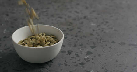 dried pumpkin seeds falling into white bowl on terrazzo countertopの写真素材