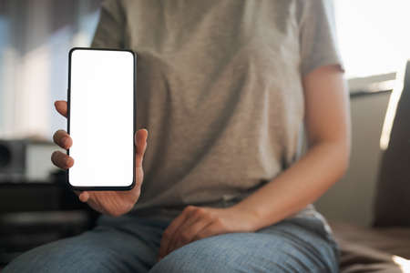Young woman show smartphone with white screen while sitting on a couch