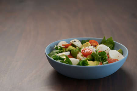 Salad with avocado, cherry tomatoes, romaine and mozzarella in blue bowl on walnut table with copy spaceの写真素材