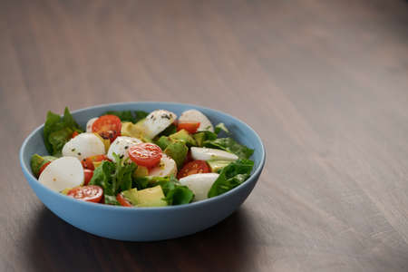 Salad with avocado, cherry tomatoes, romaine and mozzarella in blue bowl on walnut table with copy spaceの写真素材