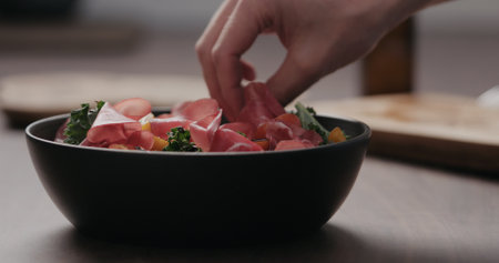 man hand add coppa slices to make salad with kale, and cherry tomatoes in black bowl on concrete...