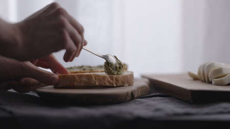 man making ciabatta bruschettas with mozzarella and pesto on olive board with window on backgorund