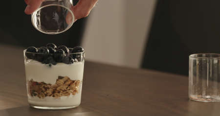 add blueberries to granola with yogurt in tumbler glass on walnut tableの写真素材