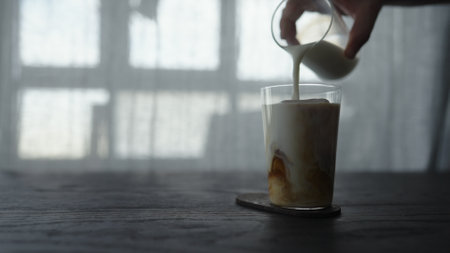 pour milk over cold brew coffee over ice cube in tumbler glass on wood tableの写真素材