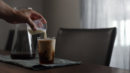 man pours milk into fresh coffee over in glass on wood table