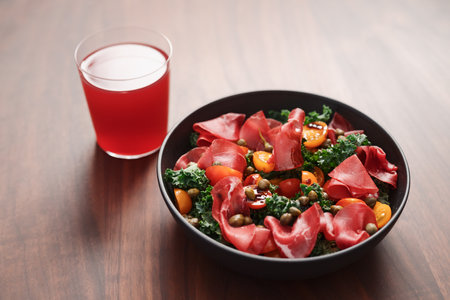 Salad with kale, bresaola and cherry tomatoes decorated with capers in black bowl and glass of red drink on walnut wood tableの写真素材