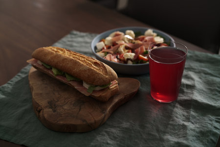 Baguette sandwich with jamon, mozzarella and salad leaves with bowl of salad and glass of lemonade for lunch on dining tableの写真素材