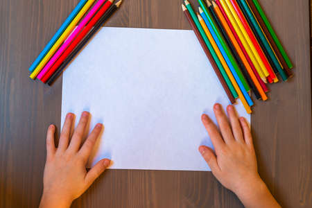The child's hands are painted with colored pencils on a white sheet of paper on a wooden table