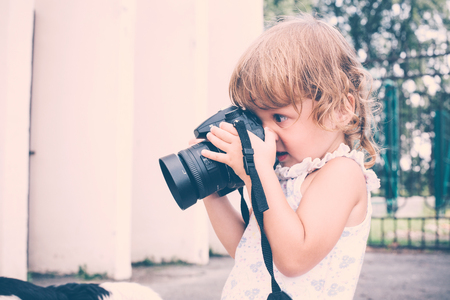 Little girl holding a camera and taking pictures. Little baby girl professional photographer outdoors. Concept development and training success.の写真素材
