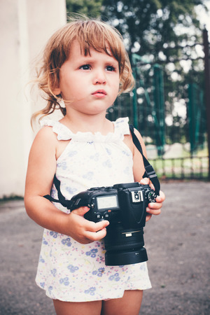 Little girl holding a camera and taking pictures. Little baby girl professional photographer outdoors. Concept development and training success.の写真素材