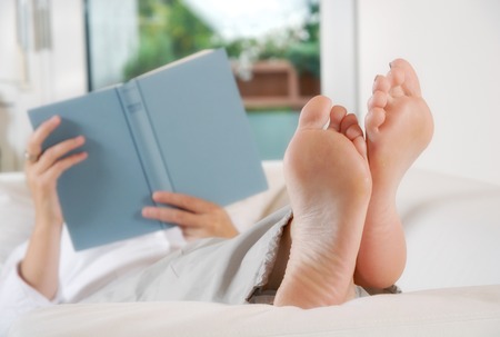 Woman lying with legs raised on a couch and reading a book の写真素材