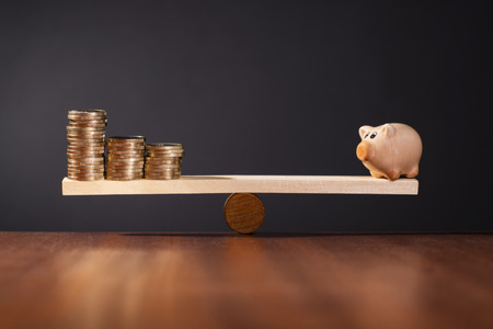Seesaw with piggy bank on one side and stack of money on the other side.の写真素材