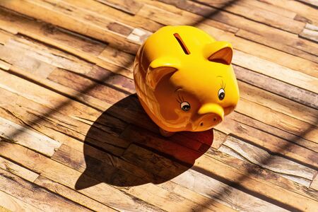 A yellow piggy bank stands on a wooden floor and casts a strong shadowの写真素材
