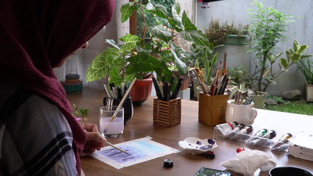 Hijab artist painting with watercolor paints on wooden table.