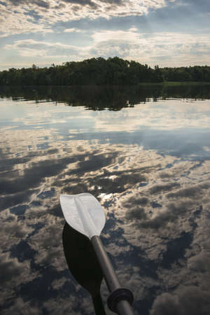 Sunrise peaking through the clouds and reflecting on the calm lake with a kayak paddle in the foregroundの写真素材