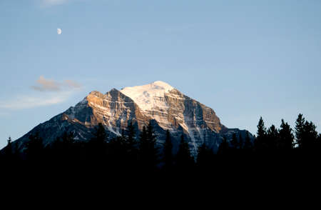 Sun hitting a rocky mountain with pine tree in the foreground and the moon in the skyの写真素材