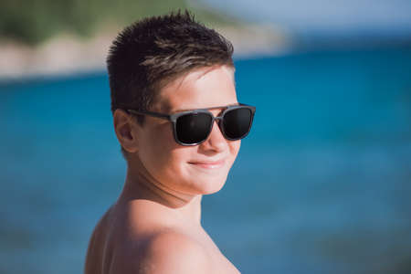 Young kid wearing sunglasses posing near the clear sea water on a beautiful summer dayの写真素材