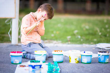 Pretty boy on the ground holding brush and painting on paper on sunny dayの写真素材