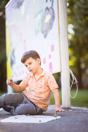 Pretty boy on the ground holding brush and painting on paper on sunny dayの写真素材