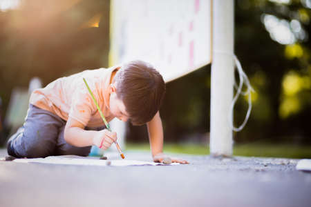 Small child playing with paintbrush on white paper into the parkの写真素材