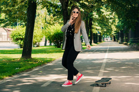 Business woman talking on cellphone in the park and driving skateboardの写真素材