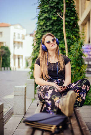 Girl listening to music streaming with headphones in summer while sitting on the benchの写真素材