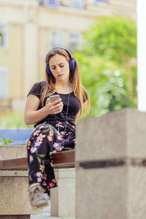 Teen girl listening to the music with headphones while sitting on the bench in the cityの写真素材