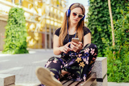Urban young woman enjoying in the music on the park while sitting on the benchの写真素材