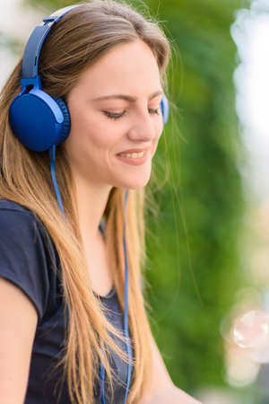 Smiling girl wearing blue headphones while sitting in the park and listening to musicの写真素材