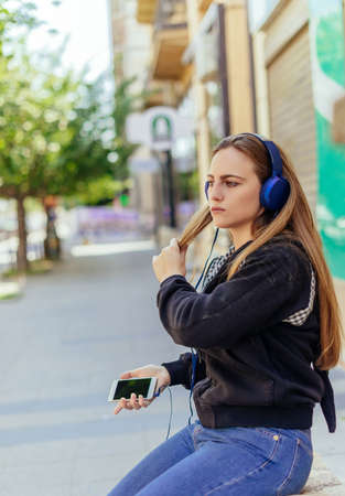 Teen girl listening to the music with headphones while sitting on the wallの写真素材
