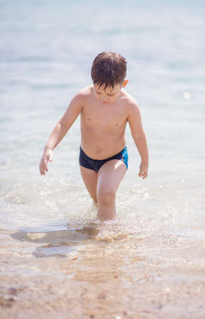 Adorable boy that wears black swimsuit is standing in the shallow water on the beachの写真素材