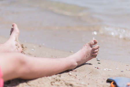 Young kid wearing red swimsuit resting his feet on the beach sand in front of the waterの写真素材