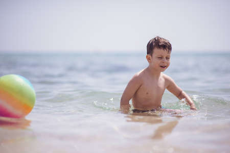 Adorable boy standing in the water on the beach holding a colorful beach ball in handsの写真素材