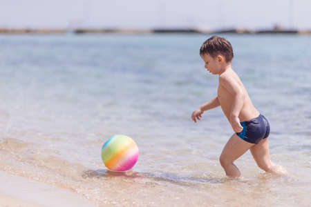 Adorable boy standing in the water on the beach playing with colorful ball and having funの写真素材