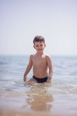 Adorable boy that wears black swimsuit is standing in the shallow water on the beachの写真素材