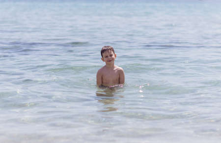 Adorable boy that wears black swimsuit is standing in the shallow water on the beachの写真素材