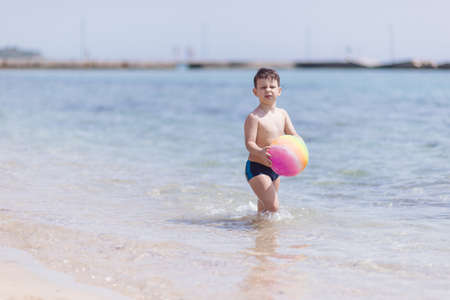 Adorable boy standing in the water on the beach holding a colorful beach ball in handsの写真素材