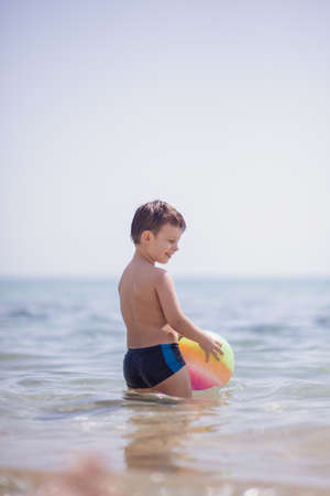Adorable boy standing in the water on the beach holding a colorful beach ball in handsの写真素材