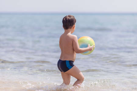 Adorable boy standing in the water on the beach holding a colorful beach ball in handsの写真素材