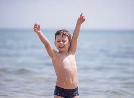Adorable boy that wears black swimsuit is standing in the shallow water on the beachの写真素材